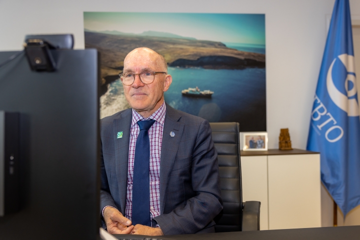 Dr Robert Floyd sits before a computer at a desk with CTBTO flag in background